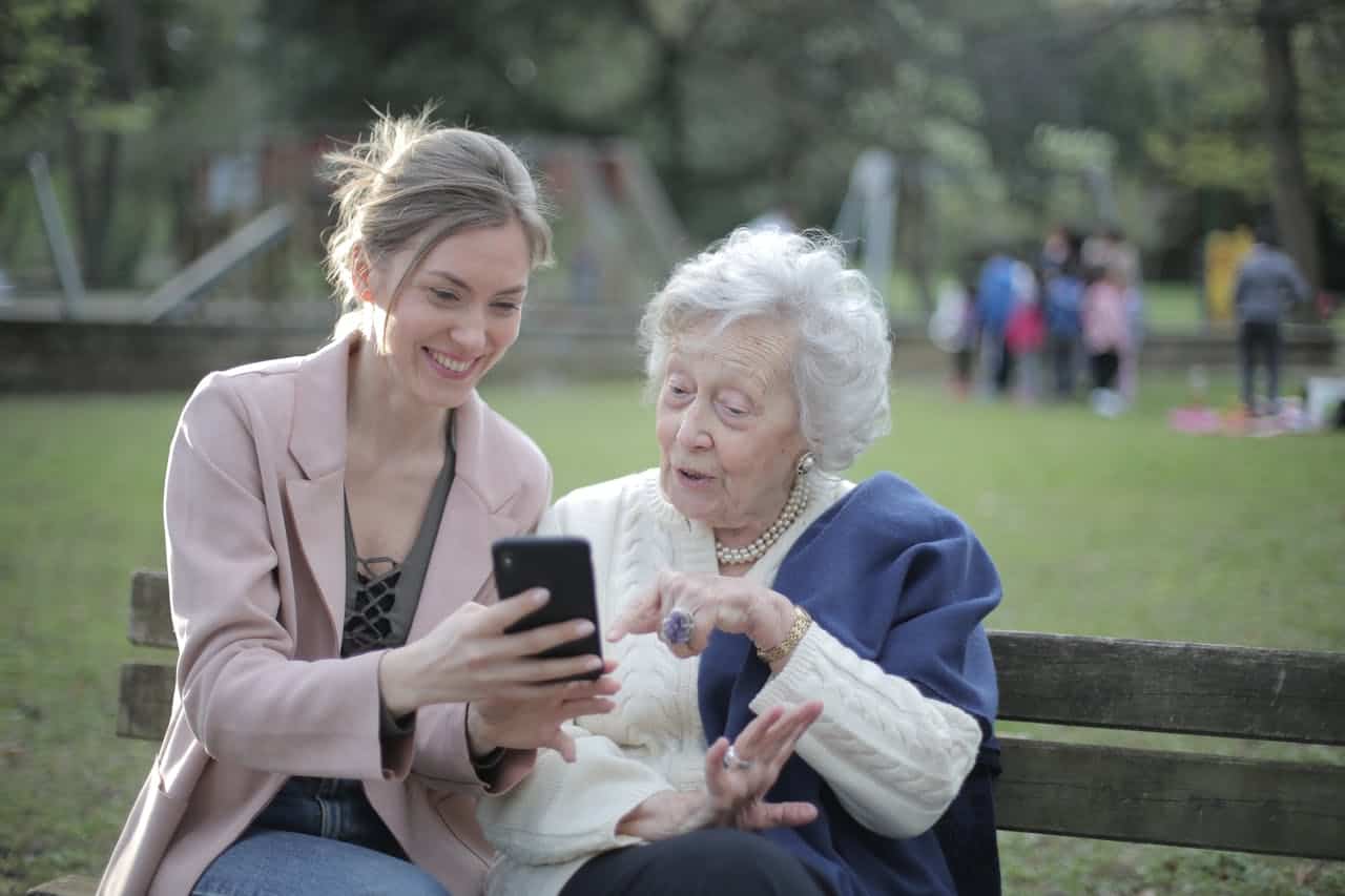 get-in-touch Delighted female relatives sitting together on wooden bench in park and browsing mobile phone while learning using